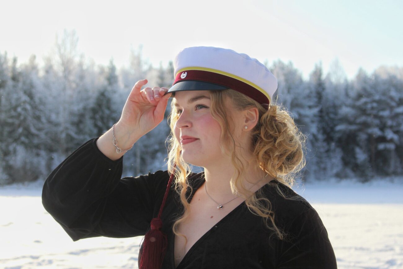 Kuvassa nainen, jolla on päässään ylioppilastyylinen lakki ja taustalla luminen maisema.