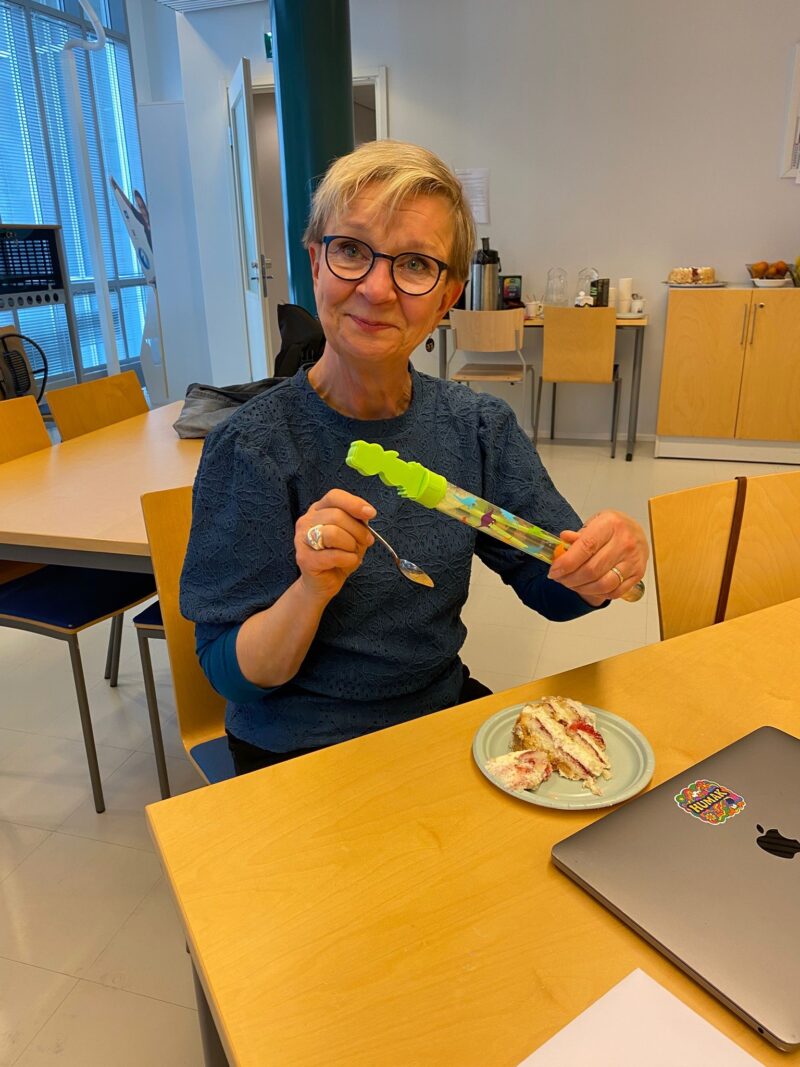 Nainen nimeltään Marjukka hymyilee kameralle kahvittelutilaisuudessa. Hänellä on toisessa kädessään lusikka ja toisessa muovinen, kirjava putkilo. Pöydällä on lautasella kakkua ja pöydällä on kannettava tietokone.