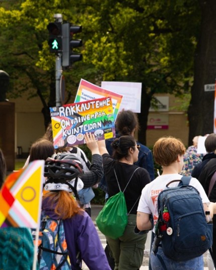 Ihmisiä Pride-kulkueessa Mikkelissä kuvattuna takaa; näkyvissä värikkäitä sateenkaarilippuja ja -kylttejä, selkiä ja reppuja, vehreä puistotausta.