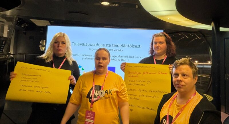 Ryhmäkuva neljästä henkilöstä. Taustalla iso näyttö,jossa lukee mustalla “tulevaisuusohjausta taidelähtöisesti”. Keskellä seisoo nuori nainen, jonka T-paidassa lukee Humak. Naisen vasemmalla puolella seisoo mustiin pukeutunut vaaleahiuksinen nainen, joka esittelee isoa keltaista kartonkia, jossa on tekstiä. Oikealla istuu mies, jolla on samanlainen T-paita kuin nuorella naisella, ja takana punahiuksinen nainen, joka esittelee isoa keltaista kartonkia. Kaikilla on kaulassa pinkki avainnauha, jossa on valkoista tekstiä.