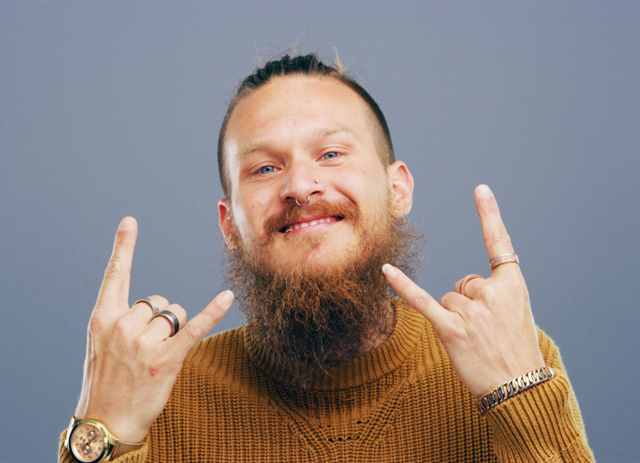Hymyilevä, parrakas mies ruskeassa neuleessa nostaa molemmat kätensä "devil horns" -sarvikäsimerkkiin, taustalla tasainen harmaa studiotausta.