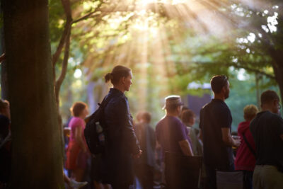 Crowd of people standing beneath trees at an outdoor music festival, watching a band as warm sunlight filters through the branches in visible rays, casting dappled light on the audience.