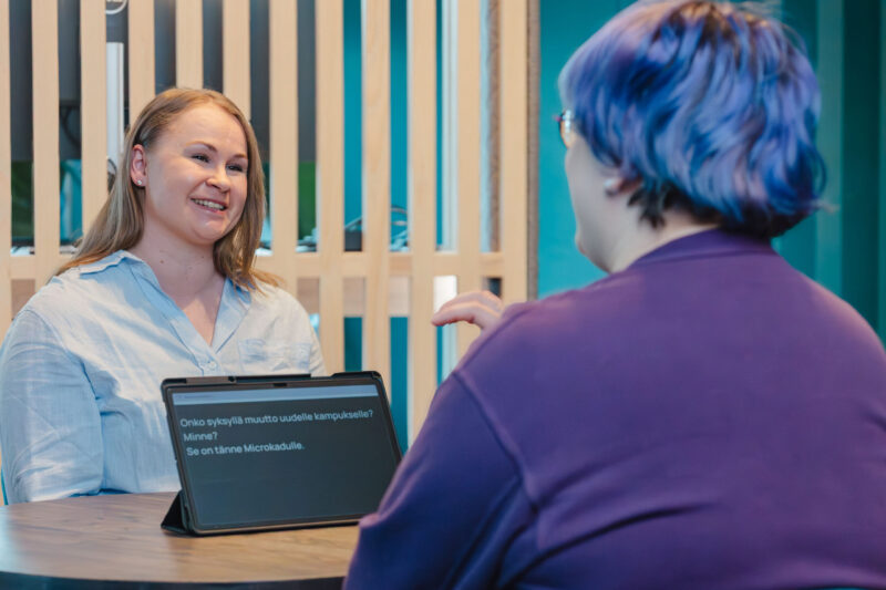 Kaksi henkilöä keskustee pöydän ääressä. Heidän välissään on tabletti, johon kirjoitustulkki kirjoittaa tekstiä.