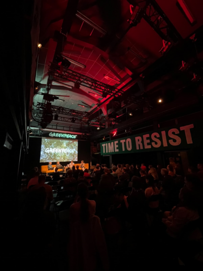 Iso sali, jossa seinällä iso banneri tekstillä "TIME TO RESIST". Salin päässä esiintymislava ja sen edessä useassa rivissä katsojia istumassa.