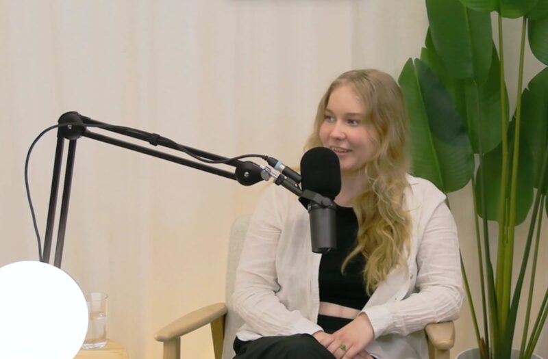 Nuori nainen istuu nojatuolissa ja puhuu mikrofoniin podcast-tyylisessä haastattelussa; hänellä on pitkä vaalea tukka, vaalea pusero ja musta paita, studiovalo ja suuret viherkasvin lehdet taustalla.