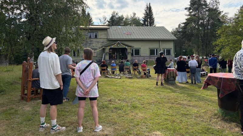 Kuvassa vanhan puutalon piha-alue, jossa seisoo ihmisiä selin kameraan seuraamssa jotain, katsee ovat suunnattu samaan suuntaan. Taustalla puita ja pilviä, ihmiset seisovat nurmikolla tai istuvat erilaisilla penkeillä.