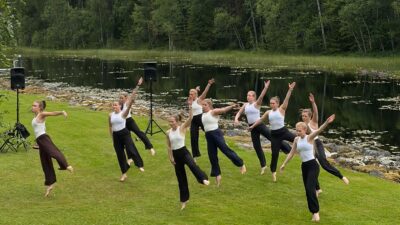 Kuvassa naisia musta-valkoisissa asusteissa ryhmävoimisteluesityksessä nurmikolla, taustalla kapea joki ja musiikkikaijutin.
