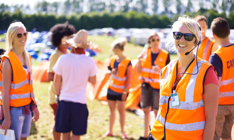 Aurinkoisella festivaalialueella oranssia heijastinliiviä käyttävä tapahtumatyöntekijä hymyilee aurinkolasit ja kuuloke‑mikrofoni päässä, muita vapaaehtoisia ja telttoja taustalla.