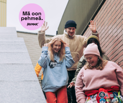 Neljä iloista nuorta laskeutumassa ulkoportaita rakennuksen vierellä; etualalla hymyilevä henkilö vaaleanpunaisessa villapaidassa ja vaaleanpunaisessa pipossa, vyöllä värikkäitä pinssejä; vieressä sininen huppari, takana beige takki ja pipo, vasemmalla osittain näkyvä keltainen takki; yläkulmassa vaaleanpunainen haalarimerkki, jossa teksti "Mä oon pehmeä."