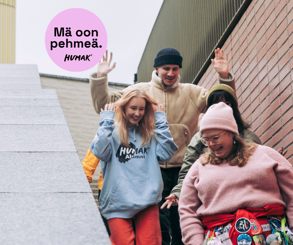 Neljä iloista nuorta laskeutumassa ulkoportaita rakennuksen vierellä; etualalla hymyilevä henkilö vaaleanpunaisessa villapaidassa ja vaaleanpunaisessa pipossa, vyöllä värikkäitä pinssejä; vieressä sininen huppari, takana beige takki ja pipo, vasemmalla osittain näkyvä keltainen takki; yläkulmassa vaaleanpunainen haalarimerkki, jossa teksti "Mä oon pehmeä."