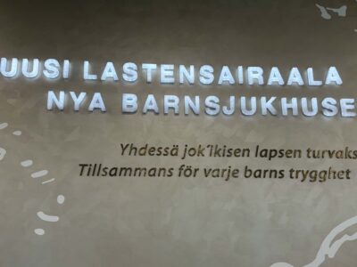 Seinäteksti: isot valaistut valkoiset kirjaimet "UUSI LASTENSAIRAALA" ja ruotsiksi "NYA BARNSJUKHUSET", alla pienemmällä "Yhdessä jok'ikisen lapsen turvaksi / Tillsammans för varje barns trygghet", vaaleabeige tausta ja valkoisia koristekuvioita.