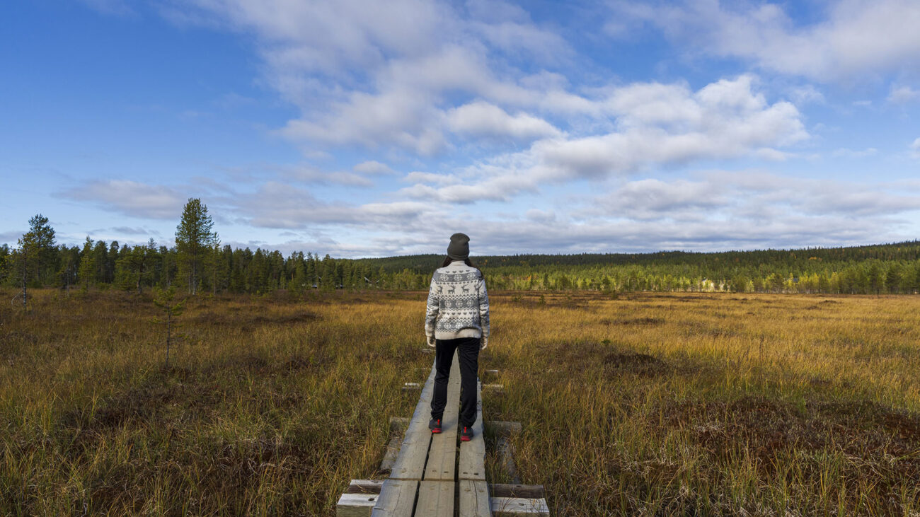Henkilö villaneuleessa ja pipossa seisoo laudoitetulla pitkospuulla keskellä avaraa suomaisemaa ja katsoo kohti horisonttia laajojen pilvien alla.