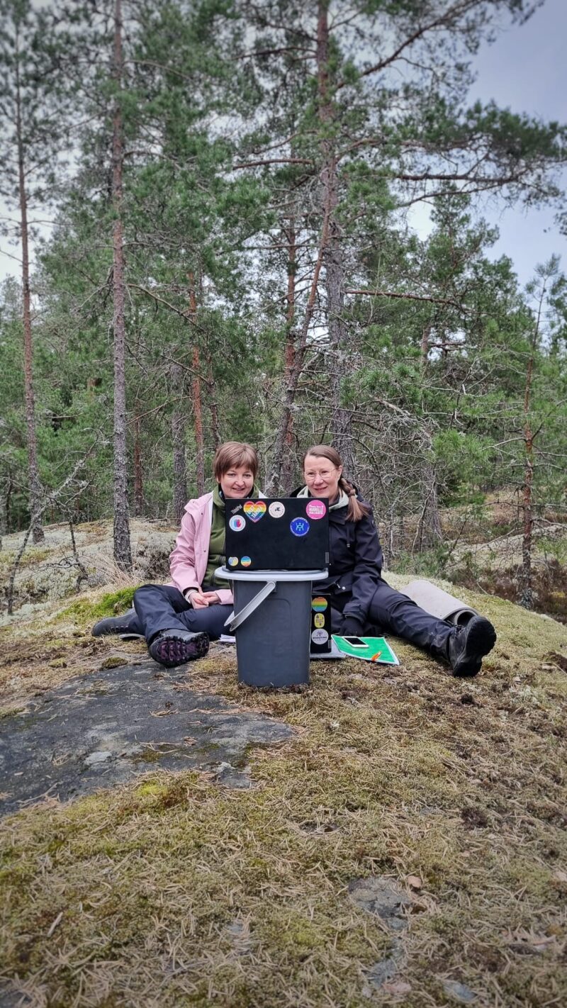 Kuvassa kaksi ihmistä istuu metsässä kalliolla kannettava tietokone edessään.