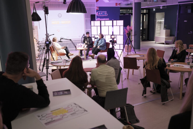 Sisäkuva Moi Cast Meet & Match -tapahtumasta Jyväskylässä: pieni yleisö istuu pöytien ja tuolien ääressä katsomassa valoisalla lavalla istuvaa paneelia, mikrofonit ja kamerat kuvaavat keskustelua ja taustalla näkyy valaistu näyttörakenne.