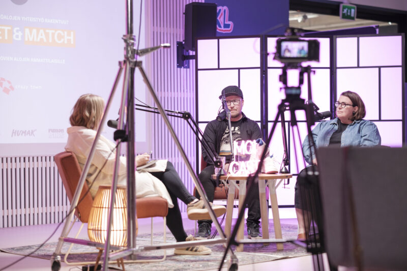 Kuva livepodcastin lavasta: kolme ihmistä istuu nojatuoleissa keskustelemassa mikrofonien ja studiokameroiden edessä, lavalla pehmeä violetti valaistus ja taustalla tapahtuman "Meet & Match" -teksti, etualalla näkyy kamera- ja valaisintelineitä.