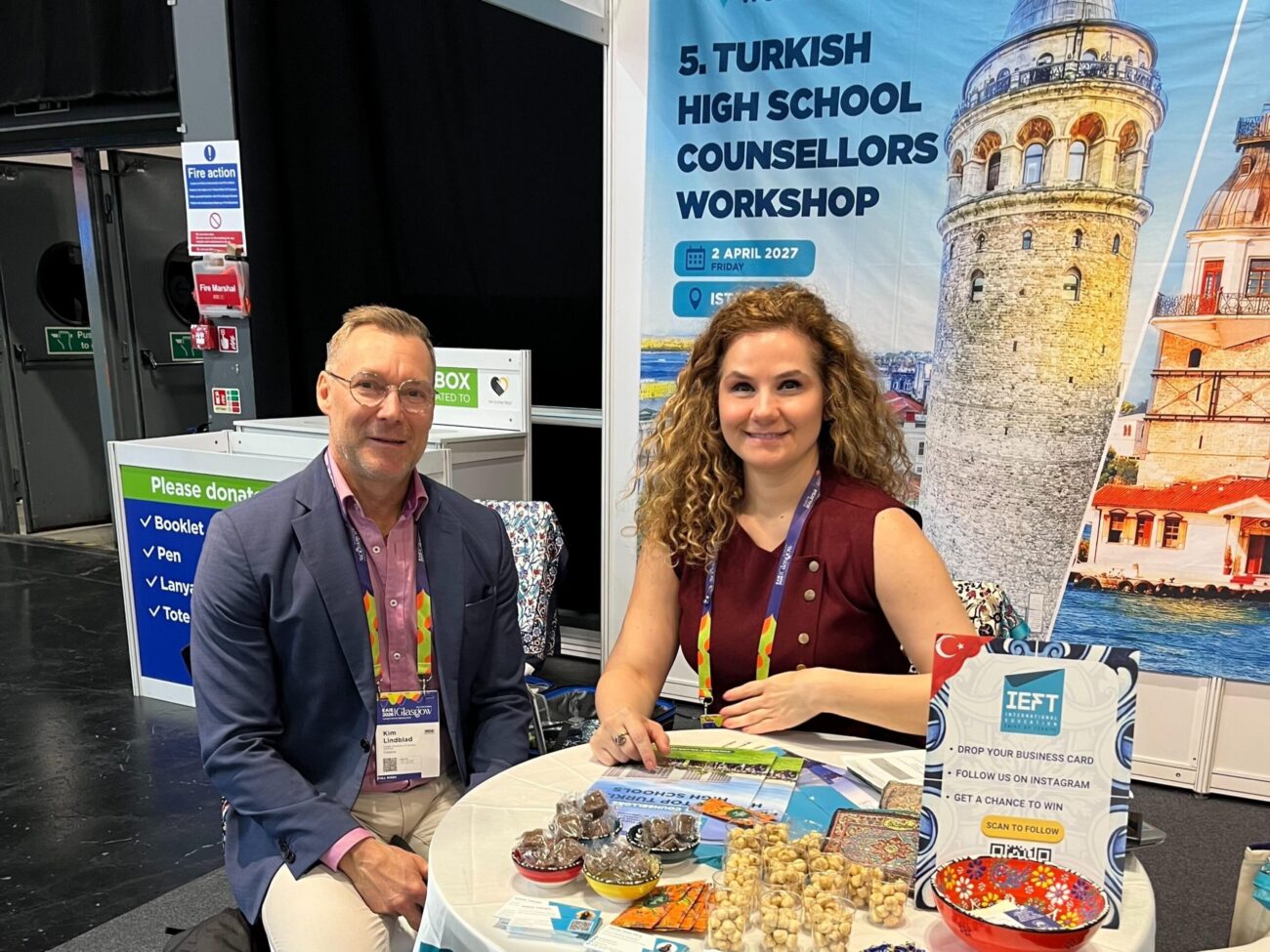Two exhibitors sit at a small round table in a trade fair booth, smiling at the camera; a man in a blazer and a woman with curly hair wear conference lanyards and have brochures, snacks and small promotional items on the table, with a large banner behind showing Istanbul’s Galata Tower and text about the Turkish High School Counsellors Workshop.