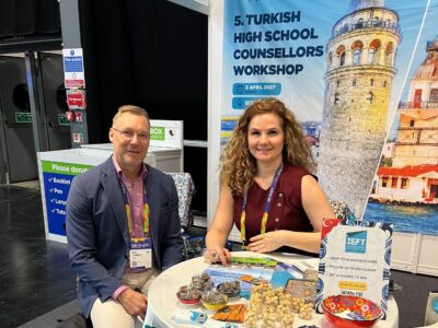 Two exhibitors sit at a small round table in a trade fair booth, smiling at the camera; a man in a blazer and a woman with curly hair wear conference lanyards and have brochures, snacks and small promotional items on the table, with a large banner behind showing Istanbul’s Galata Tower and text about the Turkish High School Counsellors Workshop.