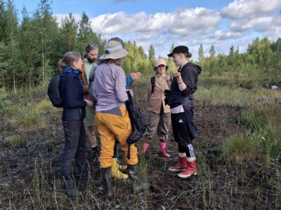 Suosymposiumin osallistujia seisomassa ja keskustelemassa Haapasuon avosuolla, ryhmä ihmisiä ulkoiluvaatteissa ja kumisaappaissa, taustalla metsäreuna ja pilvinen sininen taivas.