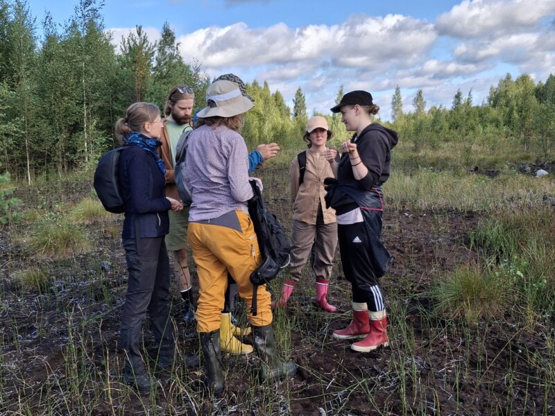 Suosymposiumin osallistujia seisomassa ja keskustelemassa Haapasuon avosuolla, ryhmä ihmisiä ulkoiluvaatteissa ja kumisaappaissa, taustalla metsäreuna ja pilvinen sininen taivas.