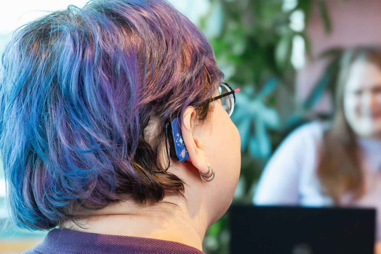 Selin kuvattu henkilö, jolla violettiin vivahtavat siniset hiukset, silmälasit ja vasemmassa korvassa sisäkorvaistute, keskustelee hymyilevän naisen kanssa pöydän toisella puolella.