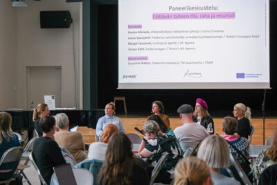 Salissa oleva yleisö katsoo heidän edessä olevaa paneelikeskustelua, yleisö on kuvan etualalla. Paneelikeskustelussa on viisi naisoletettua rivissä istumassa, rintamasuunta kohti yleisöä. Panelistien takana on valkokankaalla diaesitys, jossa lukee paneelin nimi ja panelistien nimet valkoisella pohjalla ja mustalla tekstillä.
