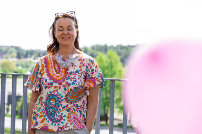 Nainen seisoo parvekkeella värikkäässä kukkakuosisessa paidassa, aurinkolasit päälaella, silmät suljettuina ja hymyillen, taustalla puustoinen maisema ja etualalla epätarkka vaaleanpunainen ilmapallo.