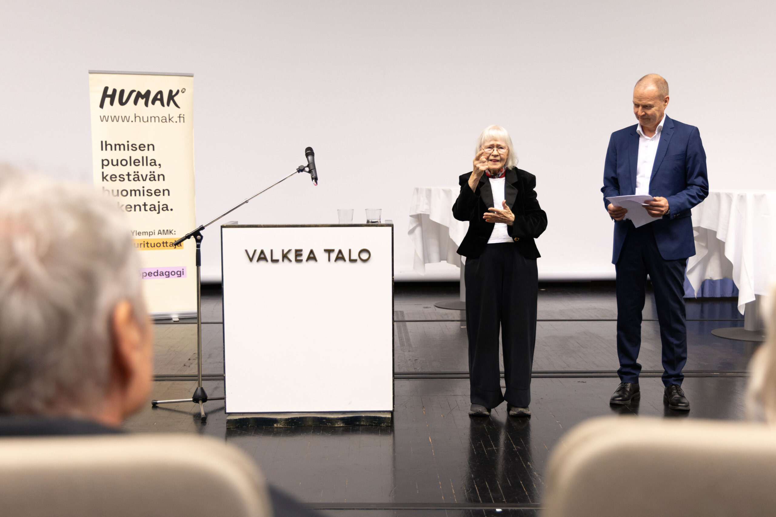 Kaksi esiintyjää seisoo lavalla valkean taustan edessä: vasemmalla iäkäs nainen valkoisin hiuksin ja mustassa puvussa hymyilee mikrofonin ääressä, oikealla mies sinisessä puvussa pitelee papereita; etualalla katsomon istuimia ja vasemmalla näkyy HUMAK-rollup sekä VALKEA TALO -pöytä.