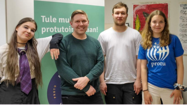 Neljä nuorta aikuista seisoo vierekkäin sisätilassa ja hymyilee kameraan; taustalla mainosjuliste, oikealla henkilö sinisessä "protu"-paidassa.
