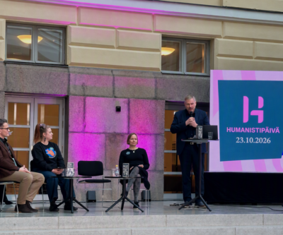 Paneelikeskustelu aulatilassa: kolme istuvaa panelistia vasemmalla ja yksi seisova puhuja puhujapöntöllä oikealla, lavalla pikkupöytiä ja vesilaseja, taustalla suuri näyttö jossa värikkäässä violetti–pinkissä valaistuksessa teksti "Humanistipäivä 23.10.2026".