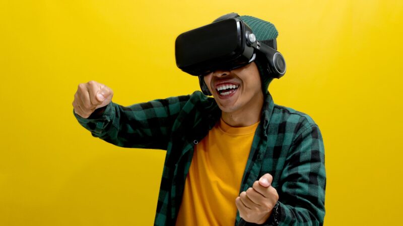 Hymyilevä henkilö käyttää virtuaalitodellisuuslaitteita ja kuulokkeita, pukeutunut vihreään ruutupaitaan ja keltaiseen t‑paitaan, kädet ojennettuina ikään kuin pelaamassa peliä kirkkaan keltaista taustaa vasten.