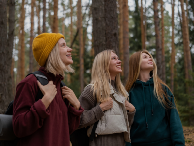 Kolme nuorta aikuista opiskelijaa metsäpolulla katsovat hymyillen ylöspäin; heillä on ulkovaatteita ja reput.