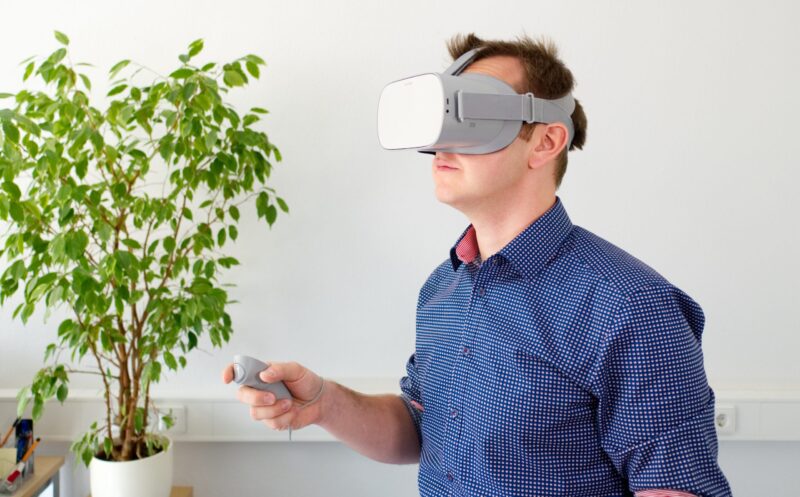 Henkilö seisoo sisätilassa ja käyttää virtuaalitodellisuuslaitteita: VR-lasit päässä ja ohjain kädessä, taustalla huonekasvi ja vaalea seinä.