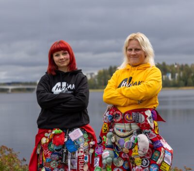 Two adult women stand side by side at a lakeshore under a cloudy sky with arms crossed and slight smiles; one has bright red hair and a black hoodie, the other has blonde hair and a yellow hoodie, and both wear colorful sashes or skirts densely covered with buttons and badges, with trees along the far shore in autumn tones.