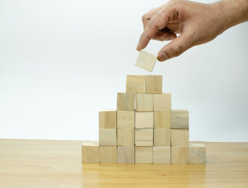 Käsi asettaa viimeistä vaaleaa puukuutiota puupalikoista muodostetun pyramidin huipulle pöydällä.