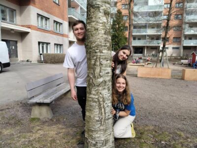 Kolme hymyilevää nuorta kurkistaa koivun takaa asuinalueen pihassa; taustalla penkki, leikkipaikka ja kerrostaloja.