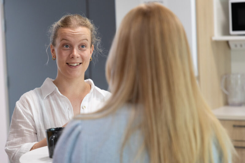 Kaksi naista istuu vastakkain pöydän ääressä keittiöhenkisessä sisätilassa; etualalla selin näkyvä vaaleatukkainen henkilö ja taustalla keskusteleva, ilmeikäs ja hymyilevä nainen.